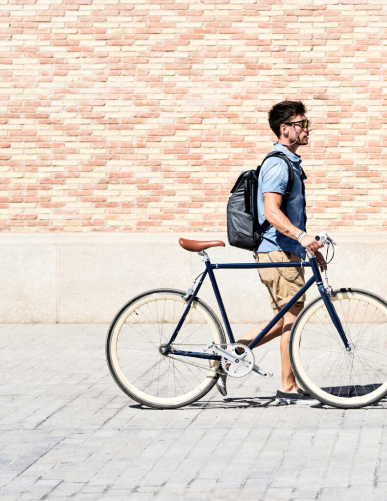 Side view of modern adult man with backpack walking down paved street with bicycle in bright sunlight