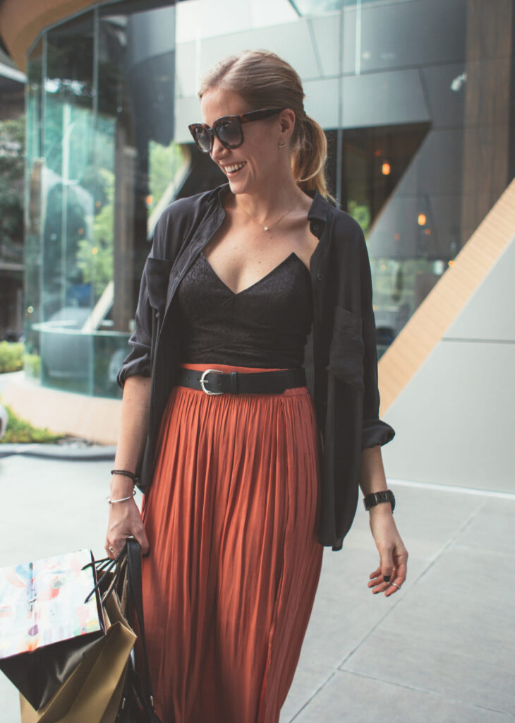 A wealthy blonde woman is smiling as she exits a mall in the city. She is wearing sunglasses and carrying shopping bags.